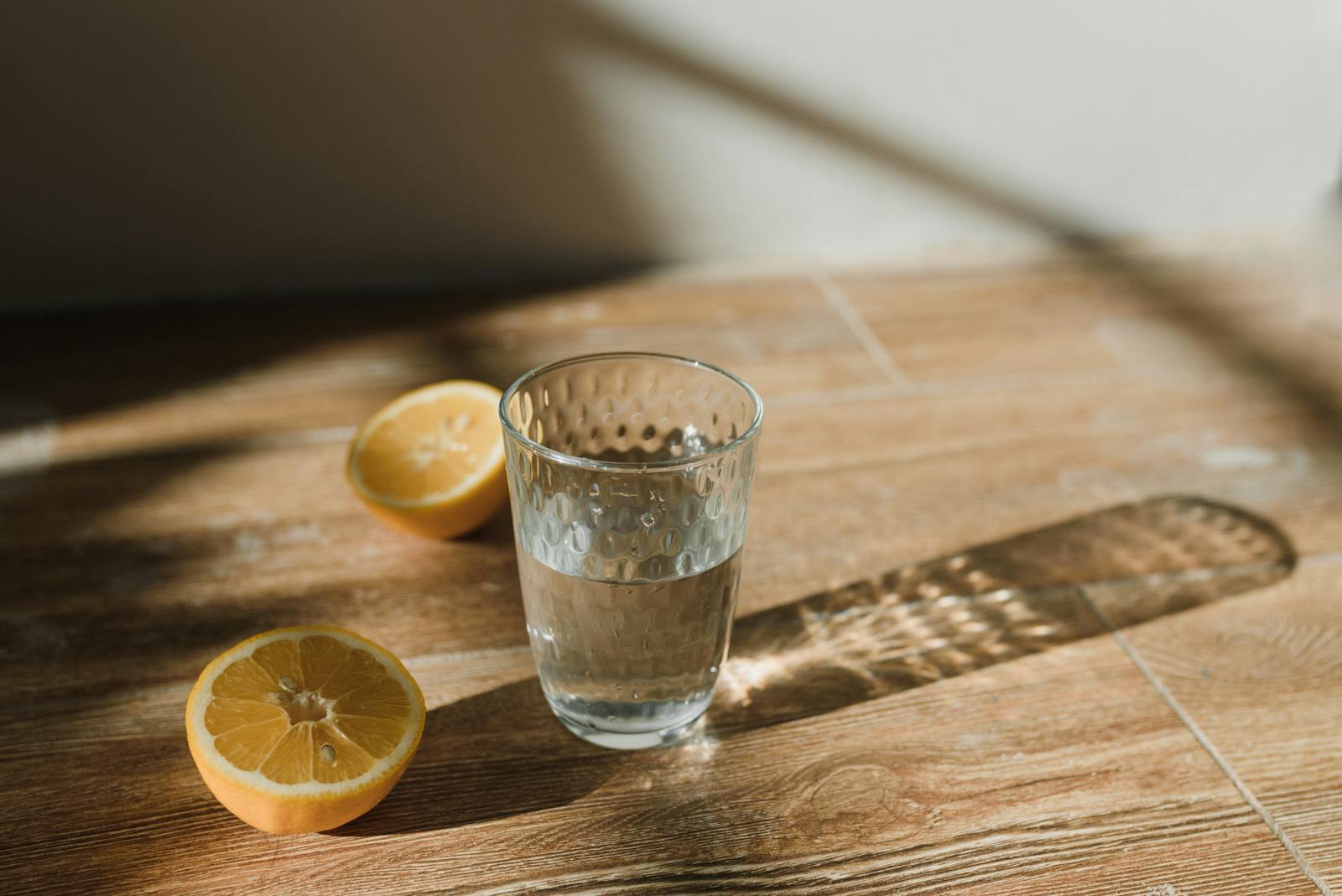Glass of distilled water beside two halved lemons on a wooden table in natural sunlight