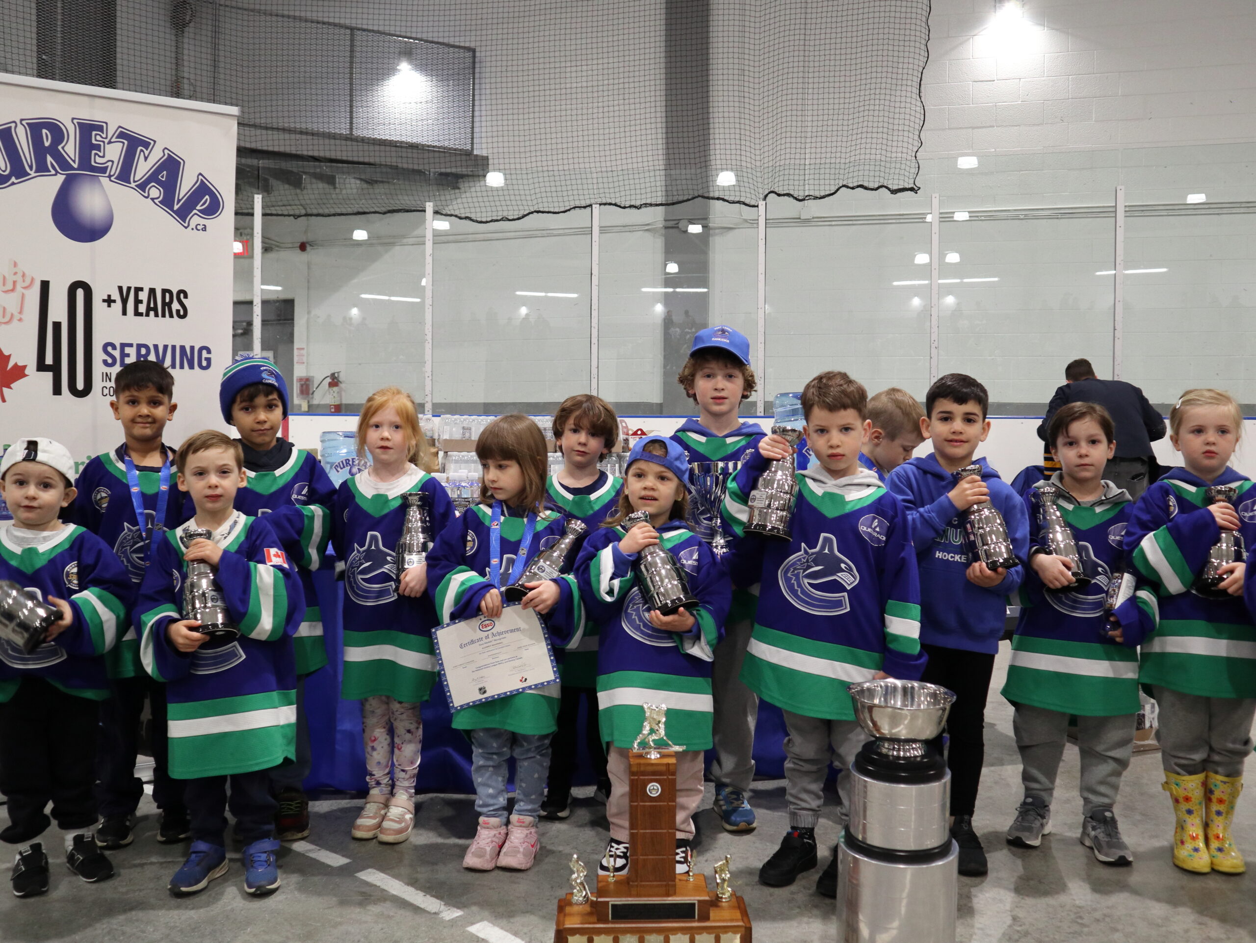 Port Credit Hockey youth team in blue, green and white jerseys posed with two large championship trophies and individual mini trophies in front of the Puretap banner at Port Credit Memorial Arena.