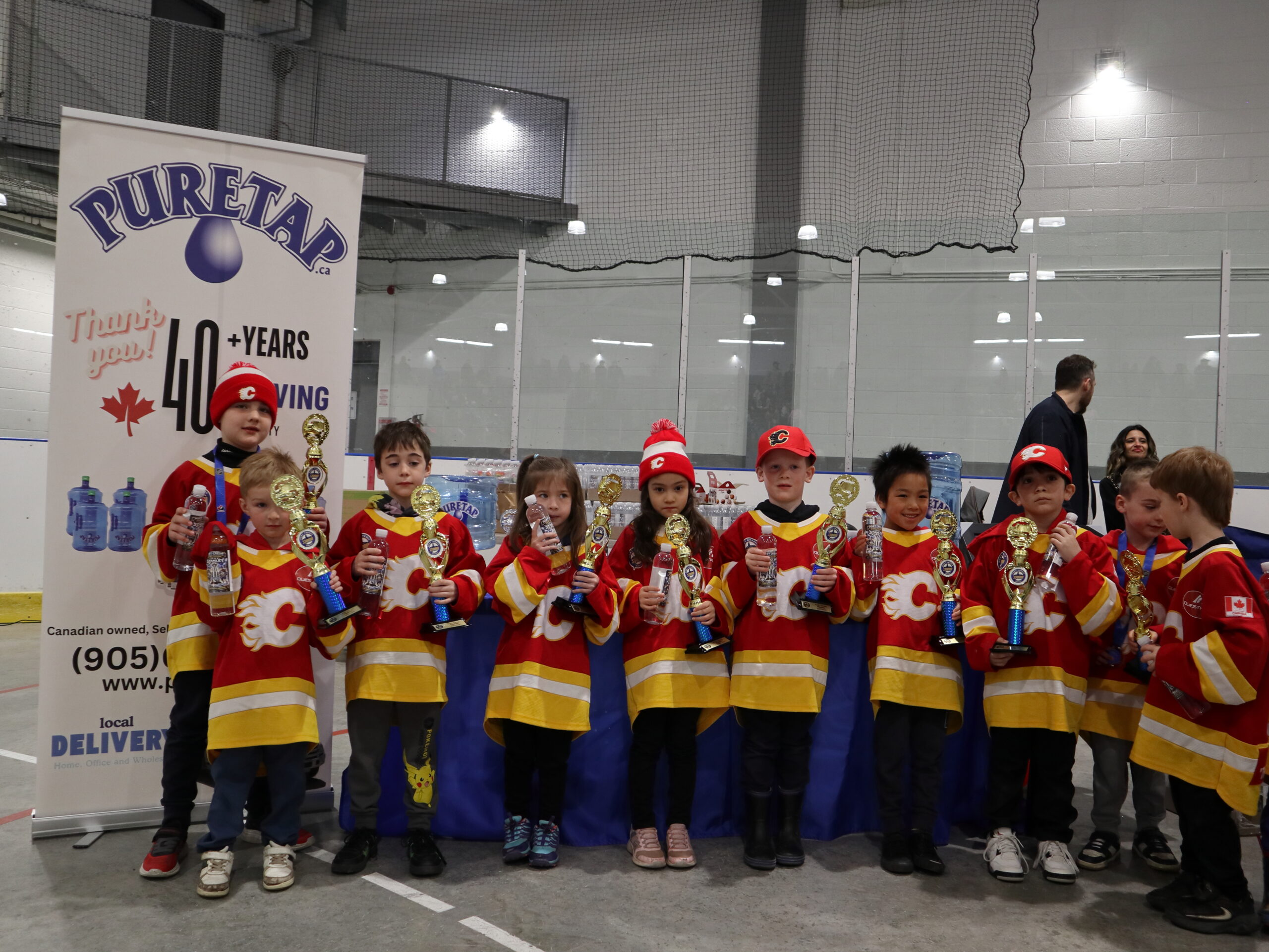Young hockey team in red and yellow jerseys holding their individual mini trophies in front of the Puretap pull-up banner at the 2026 Port Credit Hockey Awards Ceremony.