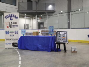 The full Puretap booth set up inside Port Credit Memorial Arena before the 2026 PCHA Awards Ceremony — two five-gallon Puretap jugs flank a table of co-branded water bottles with our pull-up banner standing behind, ready for the teams to arrive.