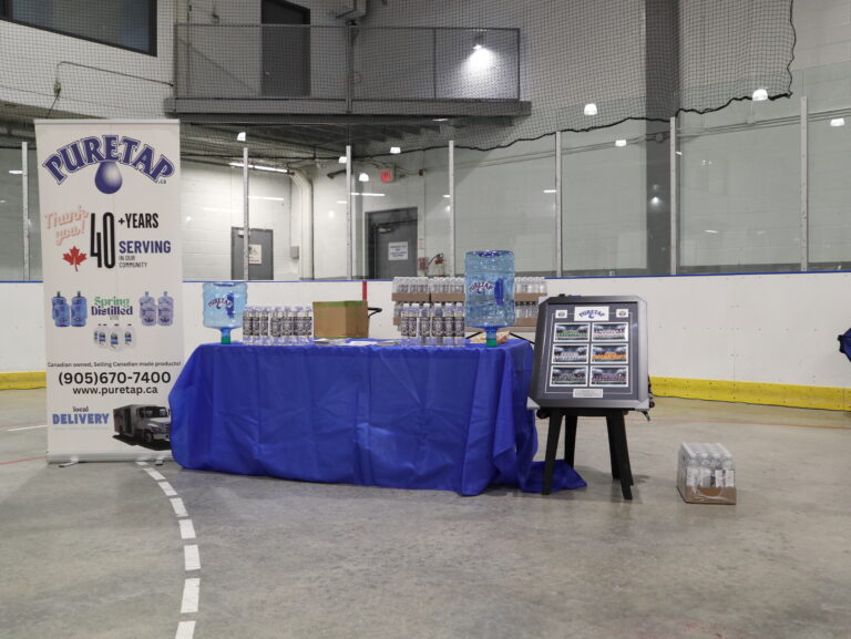 The full Puretap booth set up inside Port Credit Memorial Arena before the 2026 PCHA Awards Ceremony — two five-gallon Puretap jugs flank a table of co-branded water bottles with our pull-up banner standing behind, ready for the teams to arrive.