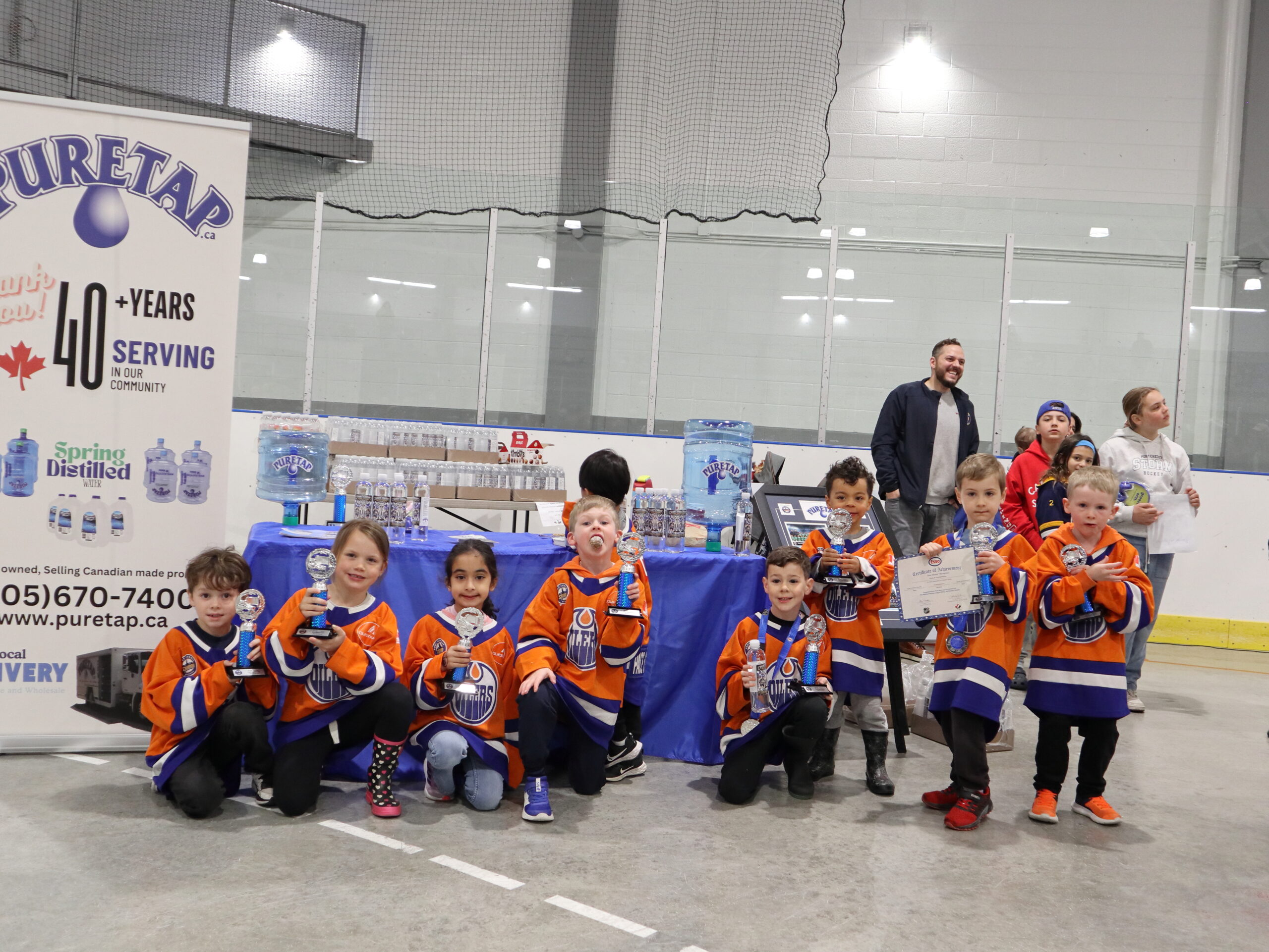 Youth hockey team in orange and blue jerseys holding their trophies in front of the Puretap booth at the 2026 Port Credit Hockey Awards Ceremony in Mississauga.