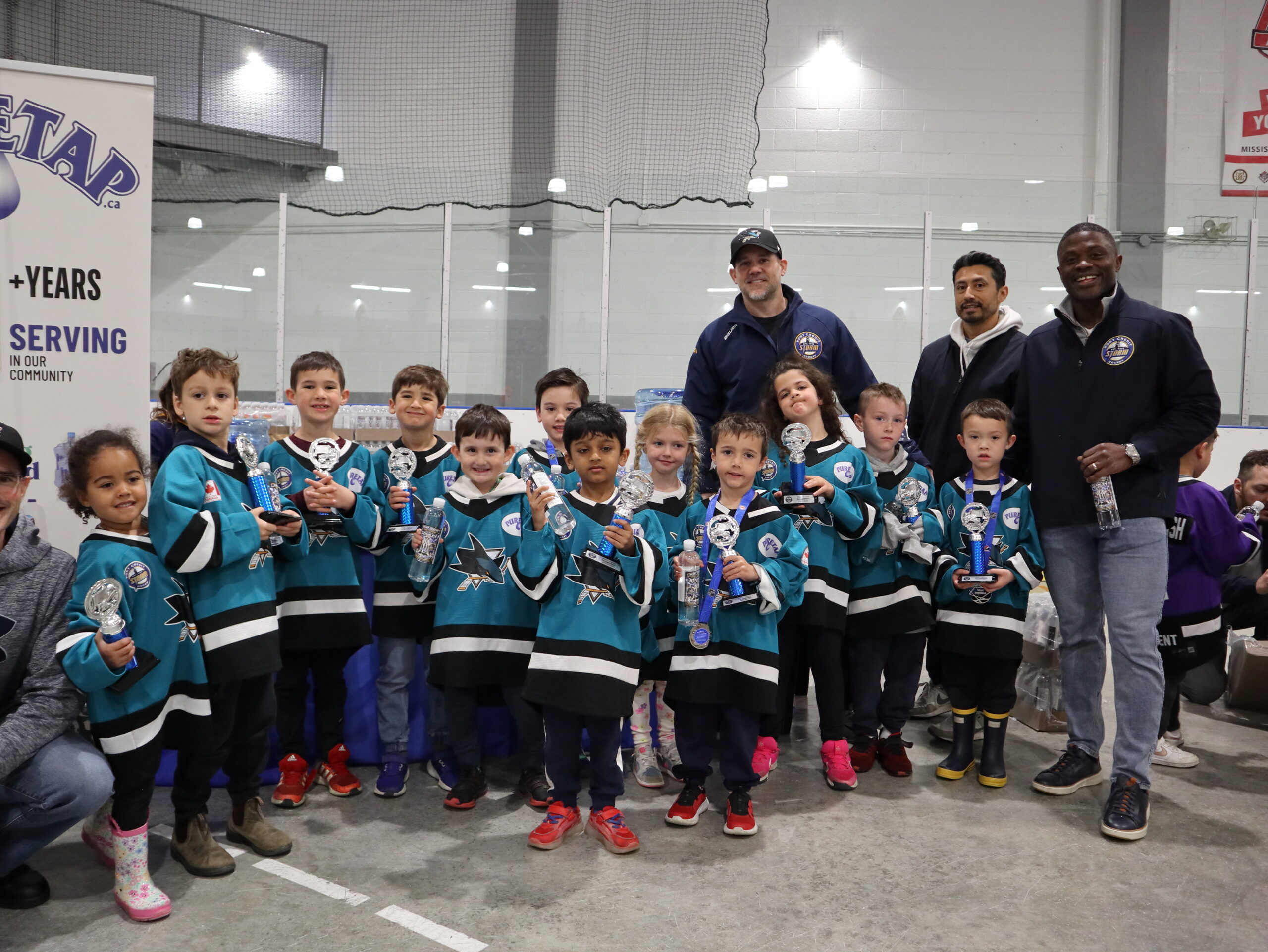 Youth hockey team in teal and black jerseys with three coaches representing diverse backgrounds, gathered with trophies and Puretap water bottles at Port Credit Memorial Arena.