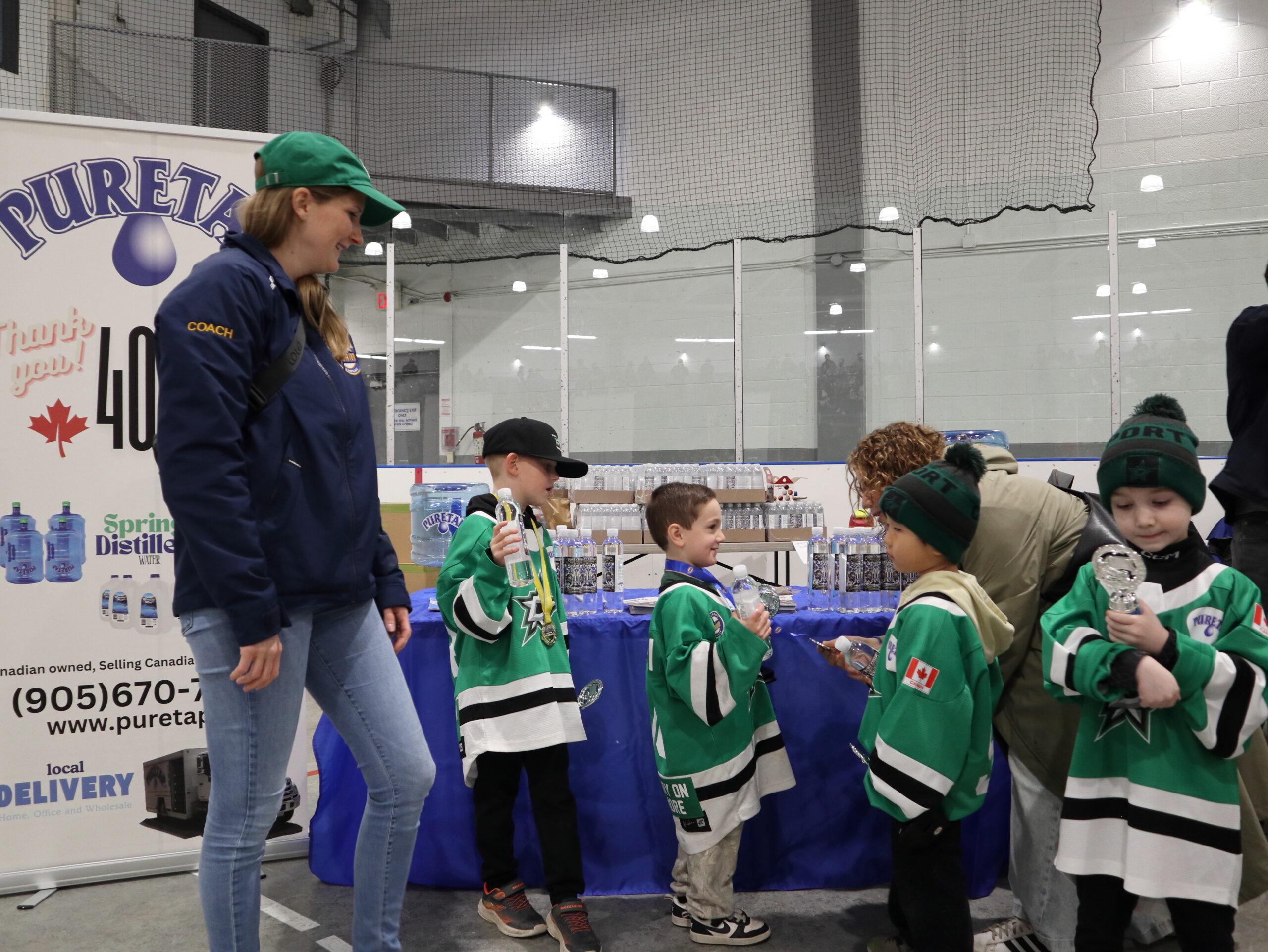 Young hockey team in green and white jerseys gathered with their female head coach at the Puretap booth, kids holding their water bottles after the awards ceremony at Port Credit Memorial Arena.