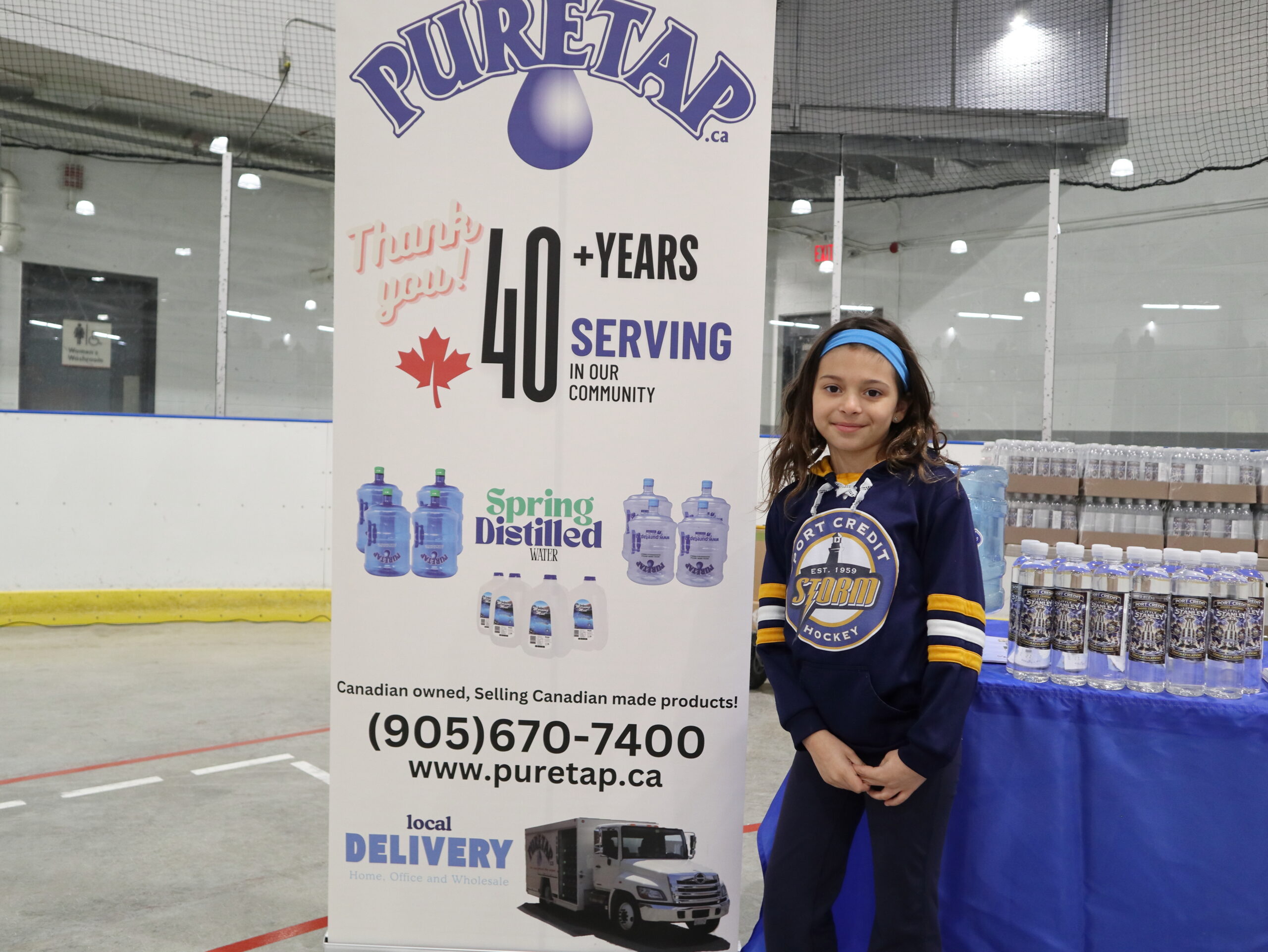 Young Port Credit Hockey player wearing a navy and yellow jersey standing beside the Puretap pull-up banner at Port Credit Memorial Arena — Puretap logo, phone number, and 40+ years tagline clearly visible.