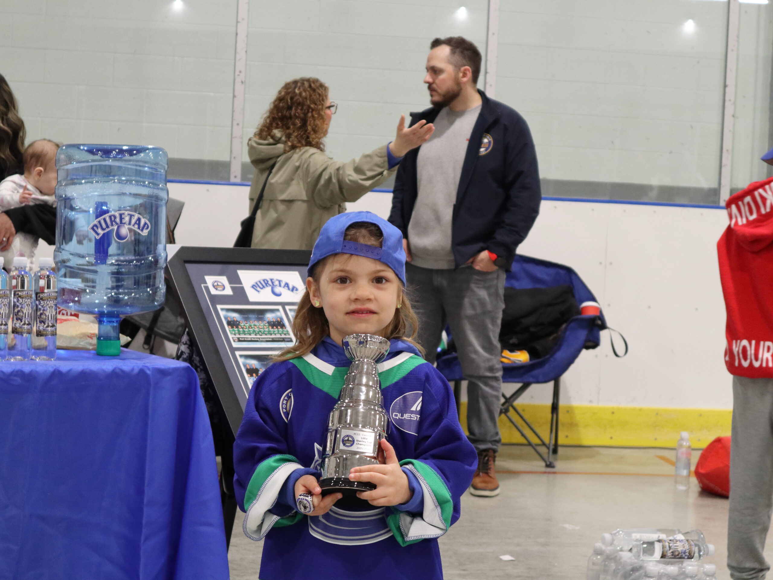Smiling young hockey player holding her individual mini trophy in front of the Puretap five-gallon jug at Port Credit Memorial Arena — a candid award-winner moment from the 2026 Port Credit Hockey Awards Ceremony.