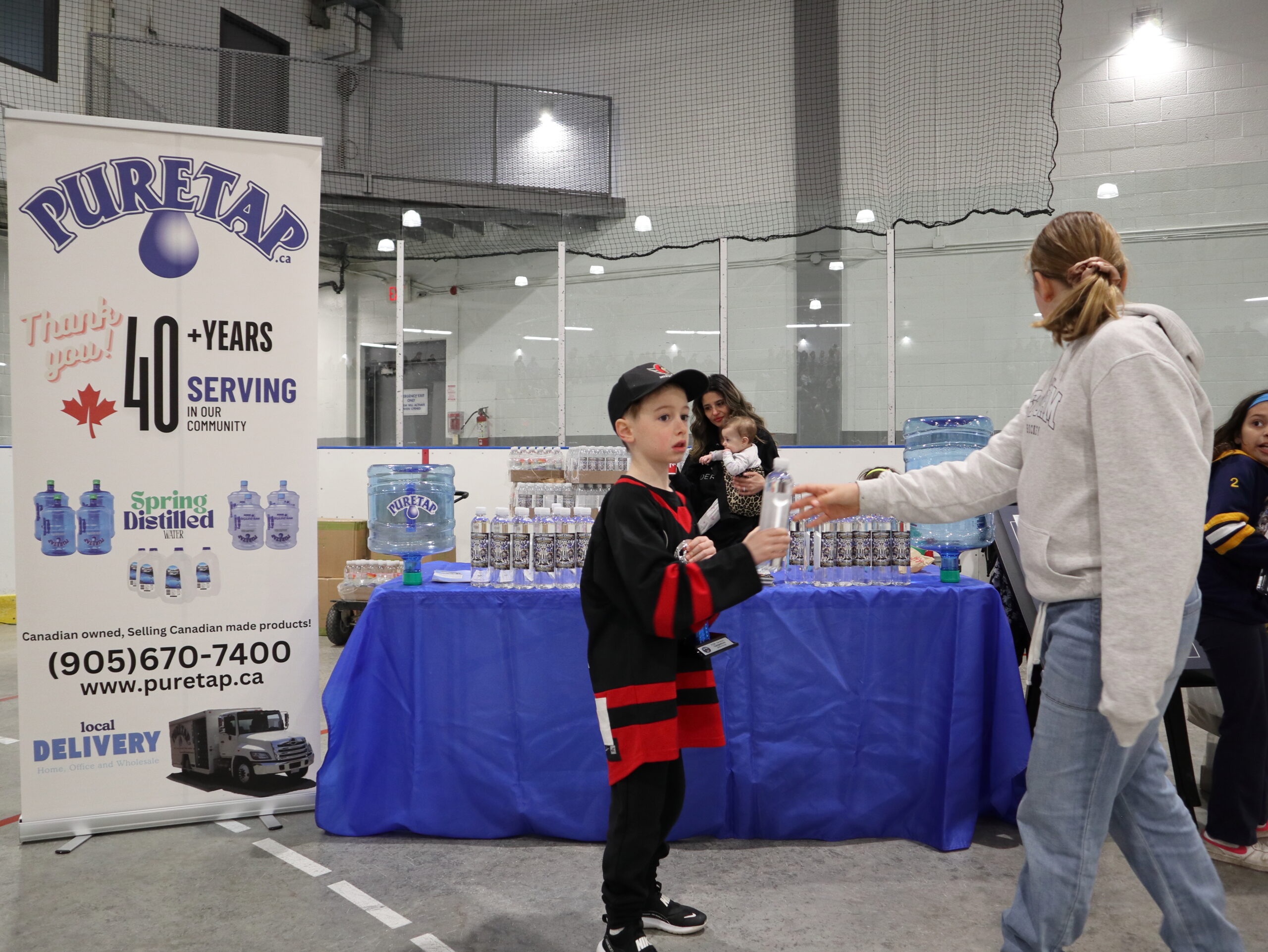 Young hockey player in a red and black jersey at the Puretap booth being handed a co-branded water bottle by a Puretap team member with the Puretap pull-up banner clearly visible.